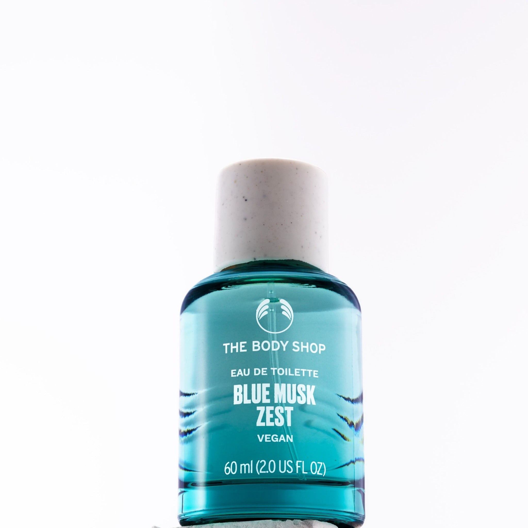 A 60ml bottle of The Body Shop Blue Musk Zest Eau de Toilette, featuring a translucent teal blue liquid and a speckled off-white cap, set against a plain white background.